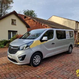 Srebrny Opel Vivaro z żółtym paskiem na tle budynku z cegły i kostki brukowej, reklama firmy przewozowej na szybie.