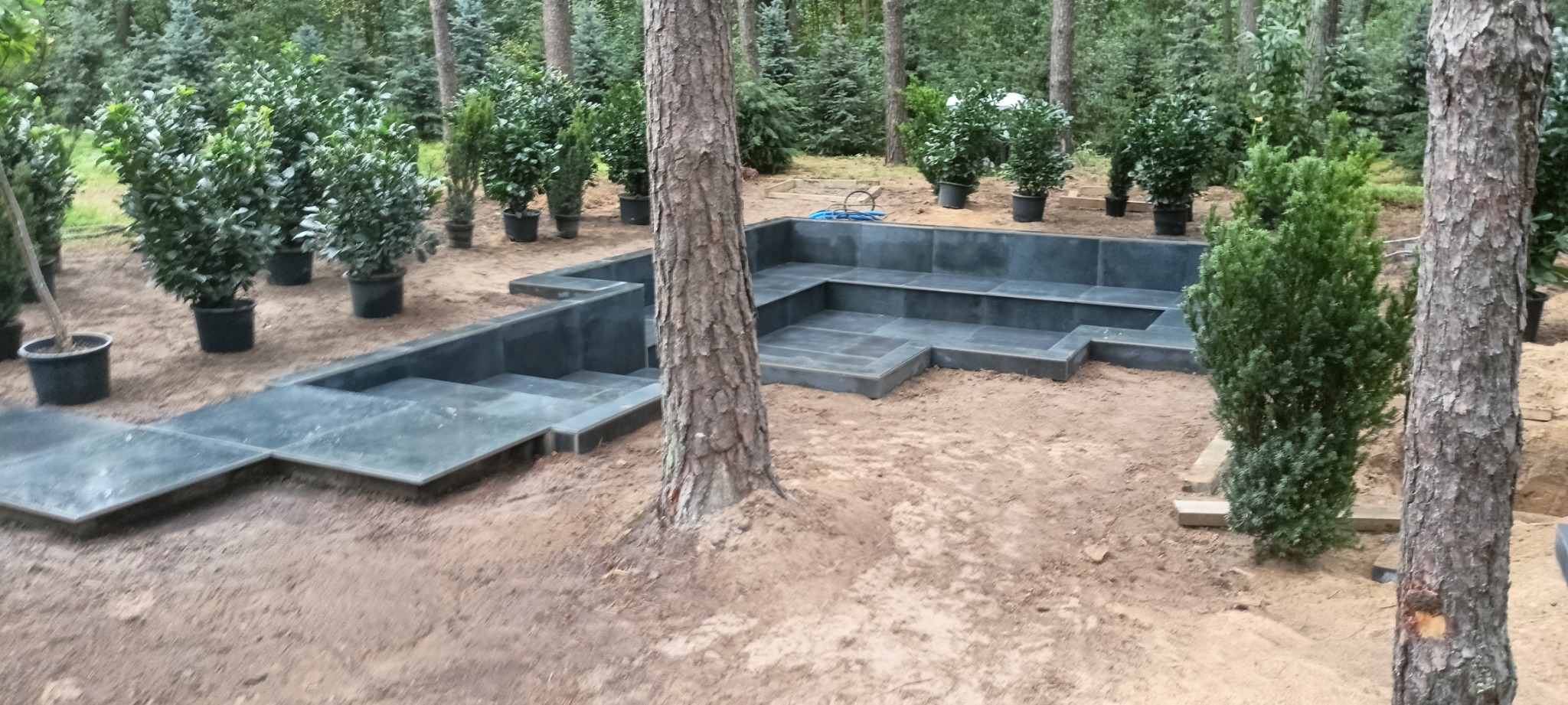 Wykończona konstrukcja z szarej kostki brukowej w ogrodzie, otoczona drzewami i roślinami w donicach, widok z perspektywy ziemi.