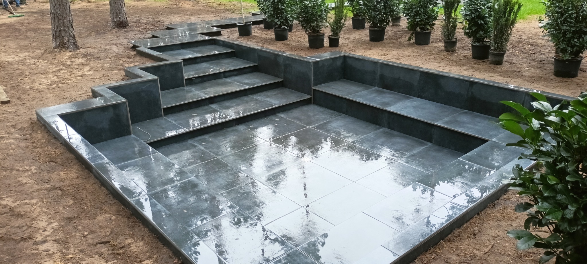 Wykończony taras zewnętrzny z ciemnoszarej, błyszczącej kostki, z niskimi ściankami, schodami i siedziskami, otoczony ziemią i roślinami w donicach.