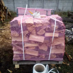 Pocięte drewno ułożone w siatce na palecie, w tle betonowy mur i stosy pni drzew