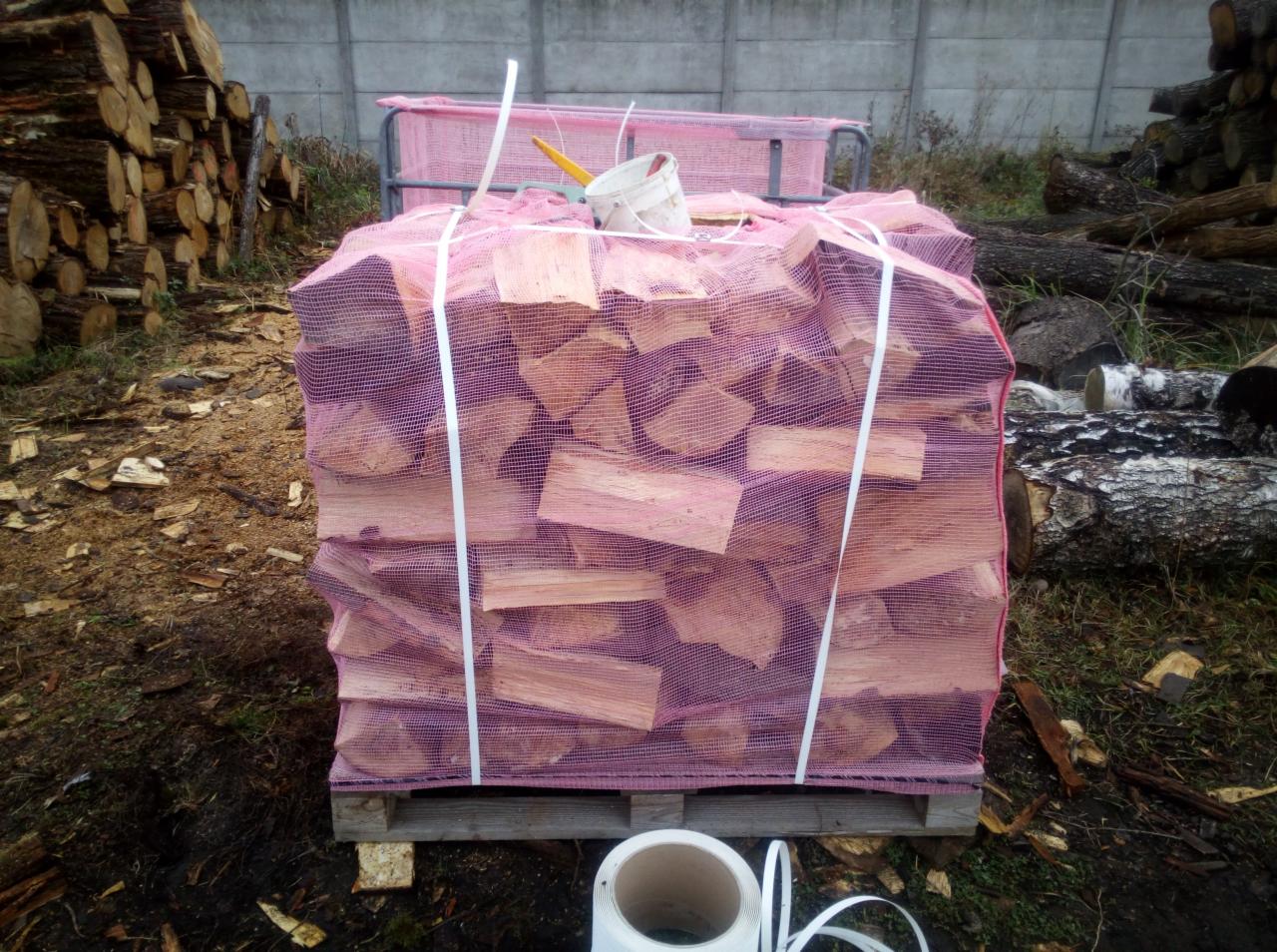 Pocięte drewno ułożone w siatce na palecie, w tle betonowy mur i stosy pni drzew