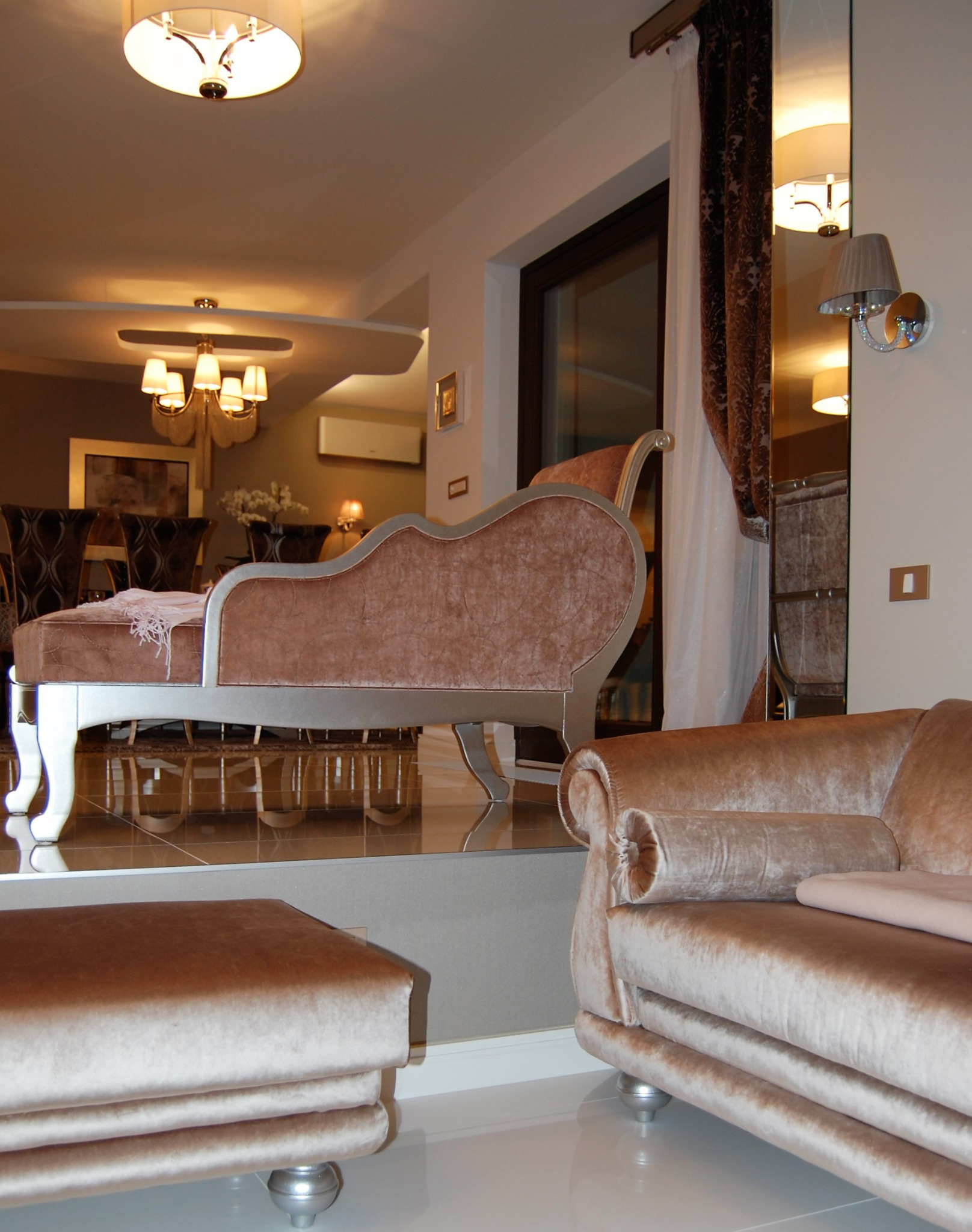 Elegancki salon z beżowymi meblami wypoczynkowymi, w tym szezlongiem i pufą, na błyszczącej podłodze, z widocznym oświetleniem sufitowym i odbiciem wnętrza w lustrze.