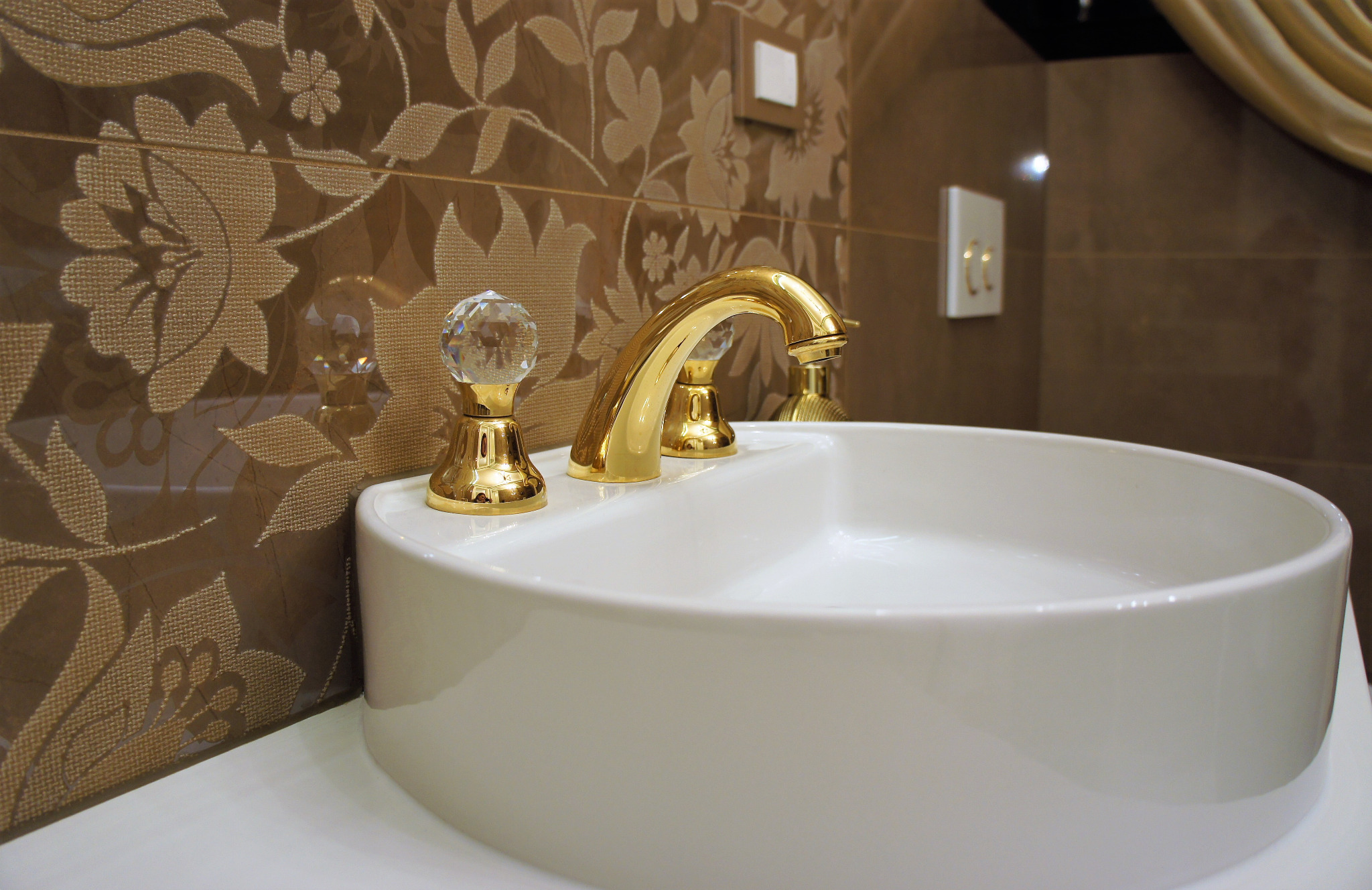 Biała, okrągła umywalka nablatowa z błyszczącą, złotą armaturą z kryształowymi uchwytami na tle brązowych płytek z kwiatowym wzorem.