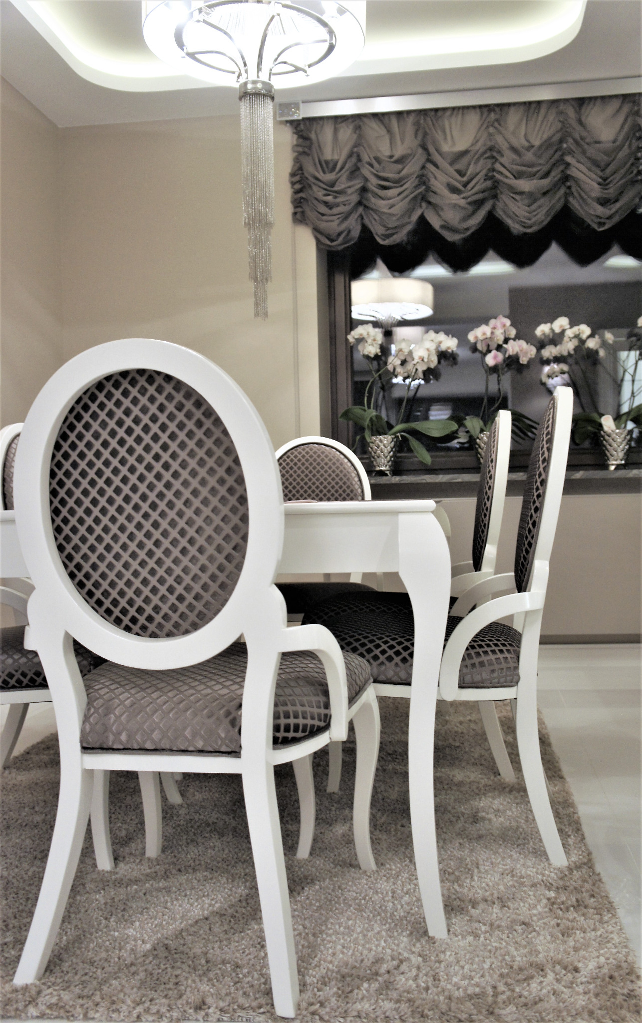 Elegancka jadalnia: biały stół z krzesłami o okrągłych oparciach i szarej tapicerce, zwisający kryształowy żyrandol, okno z orchideami i drapowaną zasłoną.