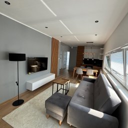 Wyrychtowane.pl - Nowoczesny salon w odcieniach szarości z drewnianymi akcentami, widok na otwartą kuchnię, oświetlenie punktowe i lampa podłogowa, jasny dywan i drewniana podłoga.