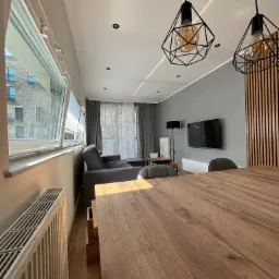 Nowoczesny salon z drewnianym stołem na pierwszym planie, szara ściana z telewizorem, geometryczne lampy wiszące i widok na budynek mieszkalny za oknem.