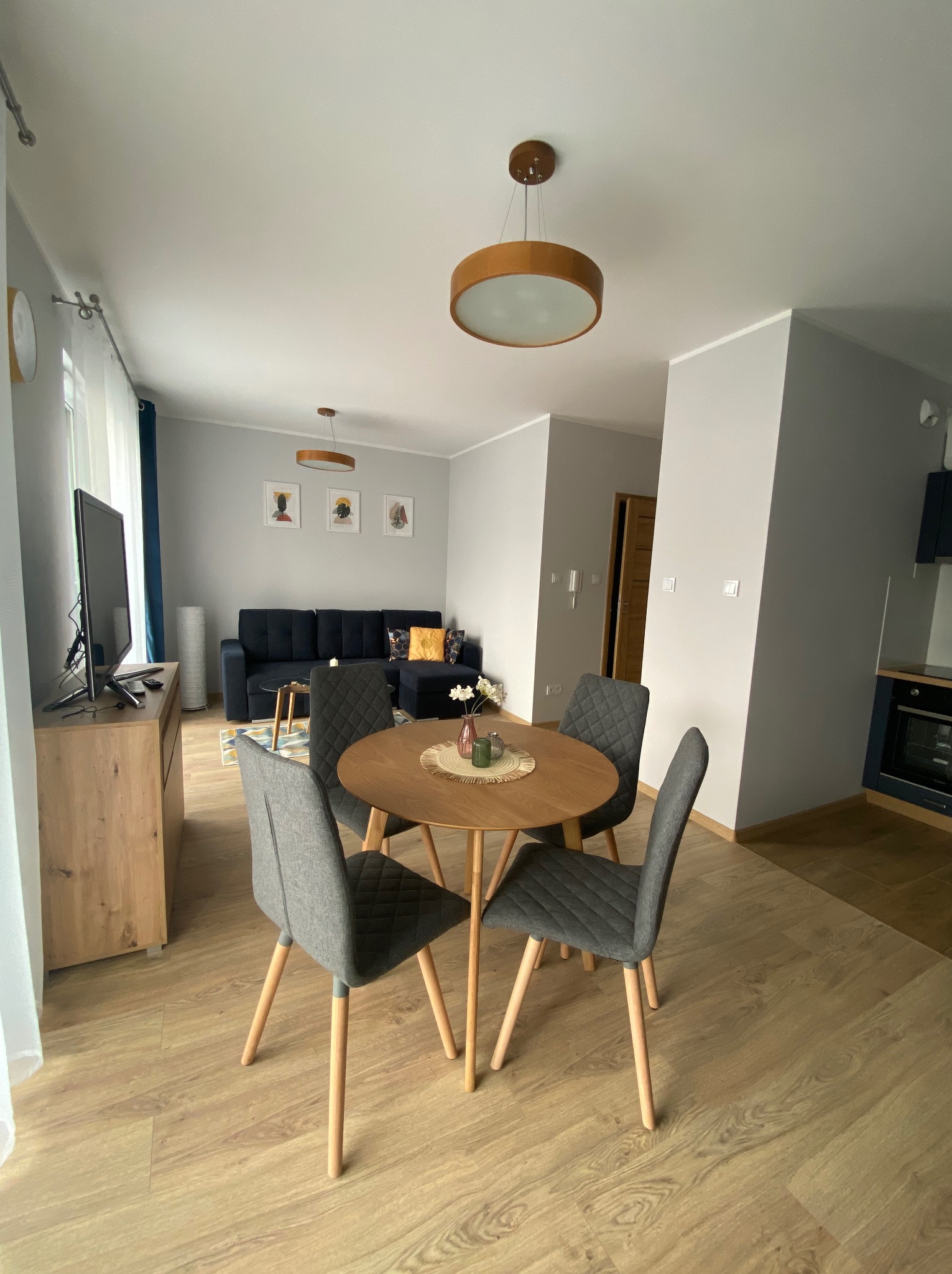 Jasne wnętrze salonu z okrągłym drewnianym stołem i czterema szarymi krzesłami na tle szarej ściany i drewnianej podłogi.