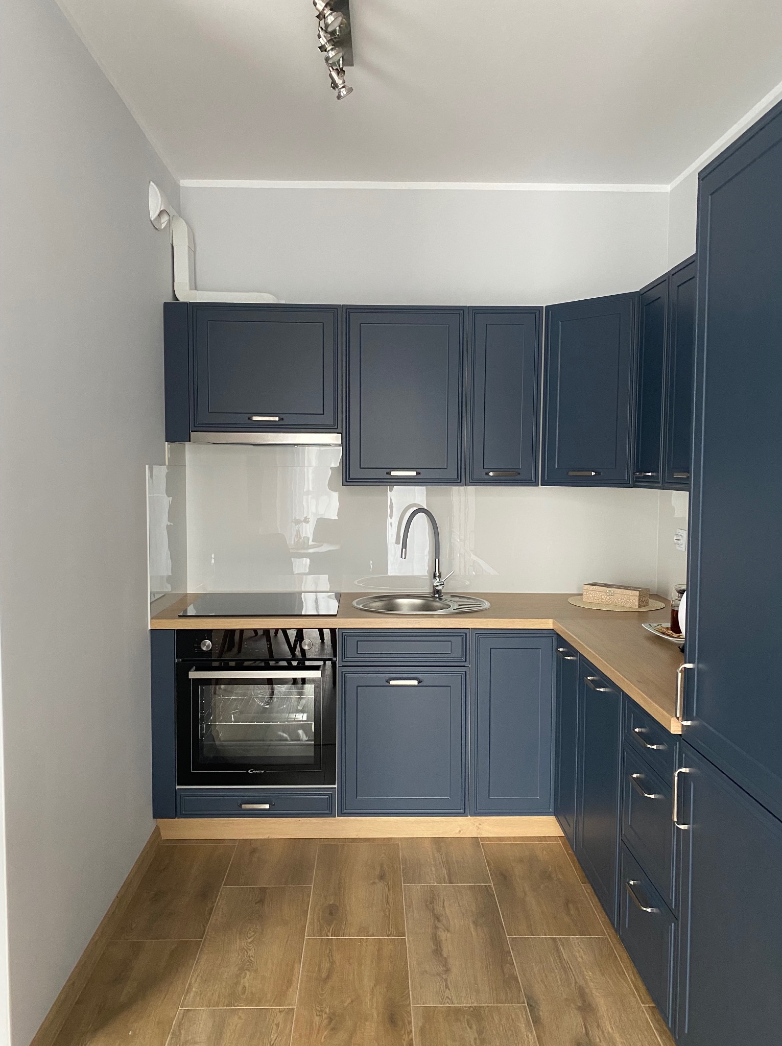 Nowoczesna kuchnia w odcieniach granatu z drewnianym blatem, ceramiczną płytą grzewczą i piekarnikiem Candy, wykończona płytkami imitującymi drewno na podłodze.