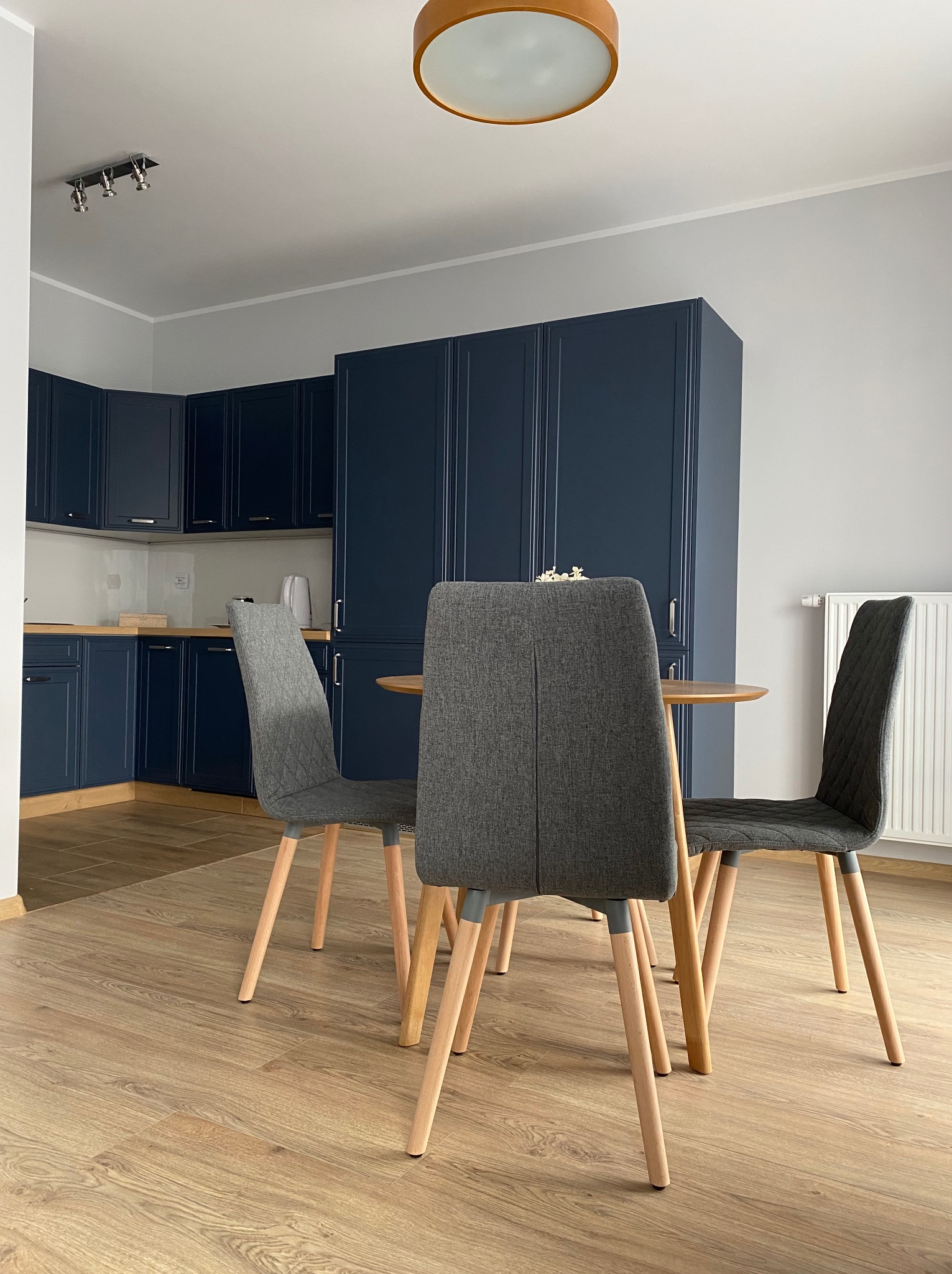 Nowoczesna kuchnia w Chorzowie z ciemnoniebieskimi szafkami, drewnianym blatem i podłogą, okrągłym stołem z trzema szarymi krzesłami i okrągłą lampą sufitową.