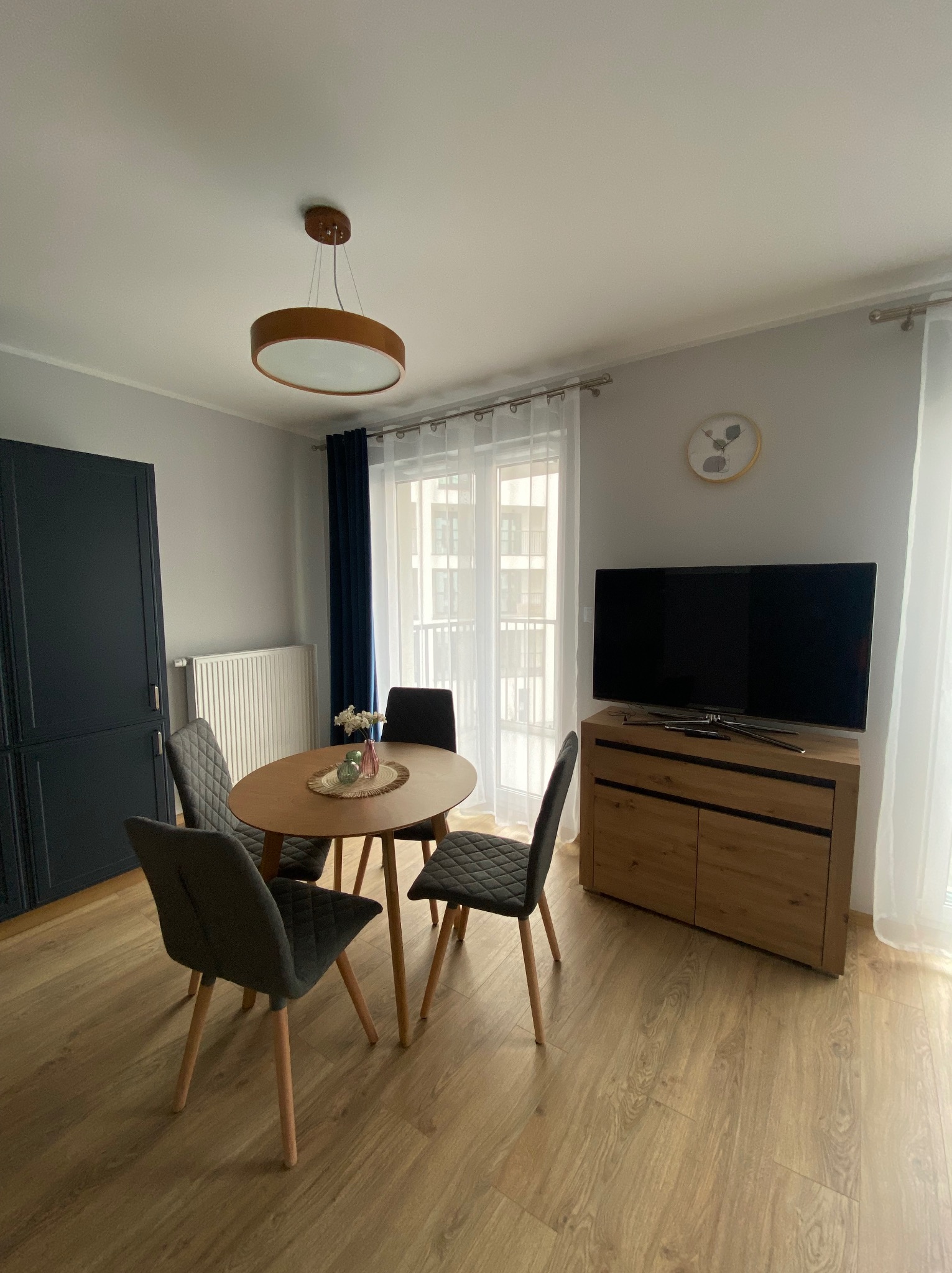 Jasne wnętrze pokoju dziennego z okrągłym stołem, czterema krzesłami z szarej tapicerki, szafką RTV pod telewizorem, ciemnogranatową szafą i białym grzejnikiem.