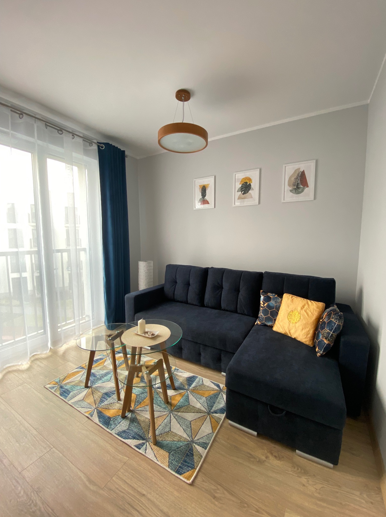 Nowoczesny salon z granatową sofą narożną, szklanym stolikiem kawowym na trójkątnym dywanie i trzema abstrakcyjnymi obrazami na szarej ścianie.