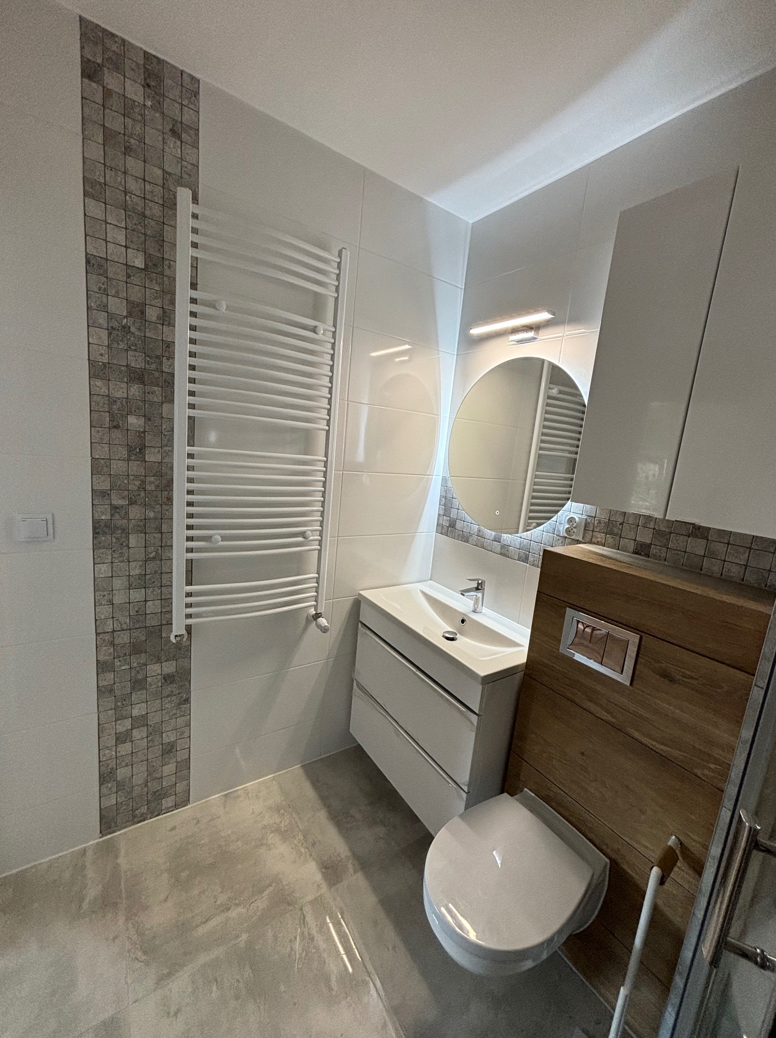 Nowoczesna łazienka z białymi płytkami, mozaiką, podwieszaną toaletą z drewnianą obudową i okrągłym lustrem z podświetleniem LED.