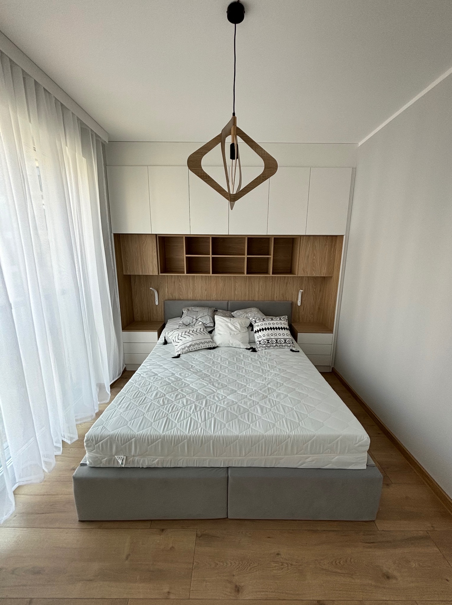 Nowoczesna sypialnia z zabudową meblową nad łóżkiem, drewnianymi półkami i designerską lampą wiszącą, doświetlona naturalnym światłem przez delikatne, białe zasłony.