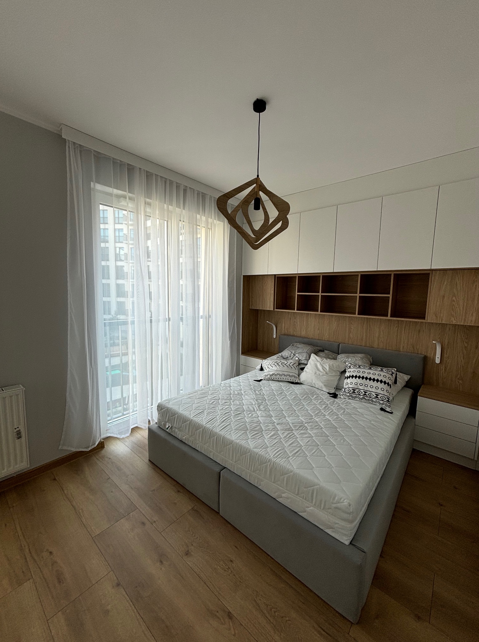 Nowoczesna sypialnia z łóżkiem kontynentalnym, drewnianą podłogą i zabudową meblową z białymi frontami oraz drewnianymi półkami, oświetlona przez okno z firanami i designerską lampę.