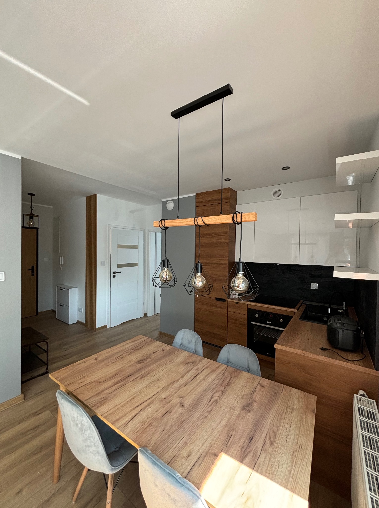 Jasne wnętrze mieszkania z drewnianym stołem, szarymi krzesłami i nowoczesnym oświetleniem nad stołem. W tle widoczna zabudowa kuchenna z elementami drewnianymi i białymi szafkami oraz fragment...