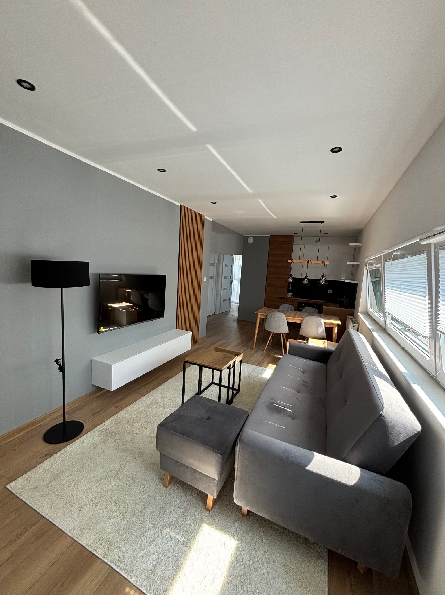 Nowoczesny salon w odcieniach szarości z drewnianymi akcentami, widok na otwartą kuchnię, oświetlenie punktowe i lampa podłogowa, jasny dywan i drewniana podłoga.