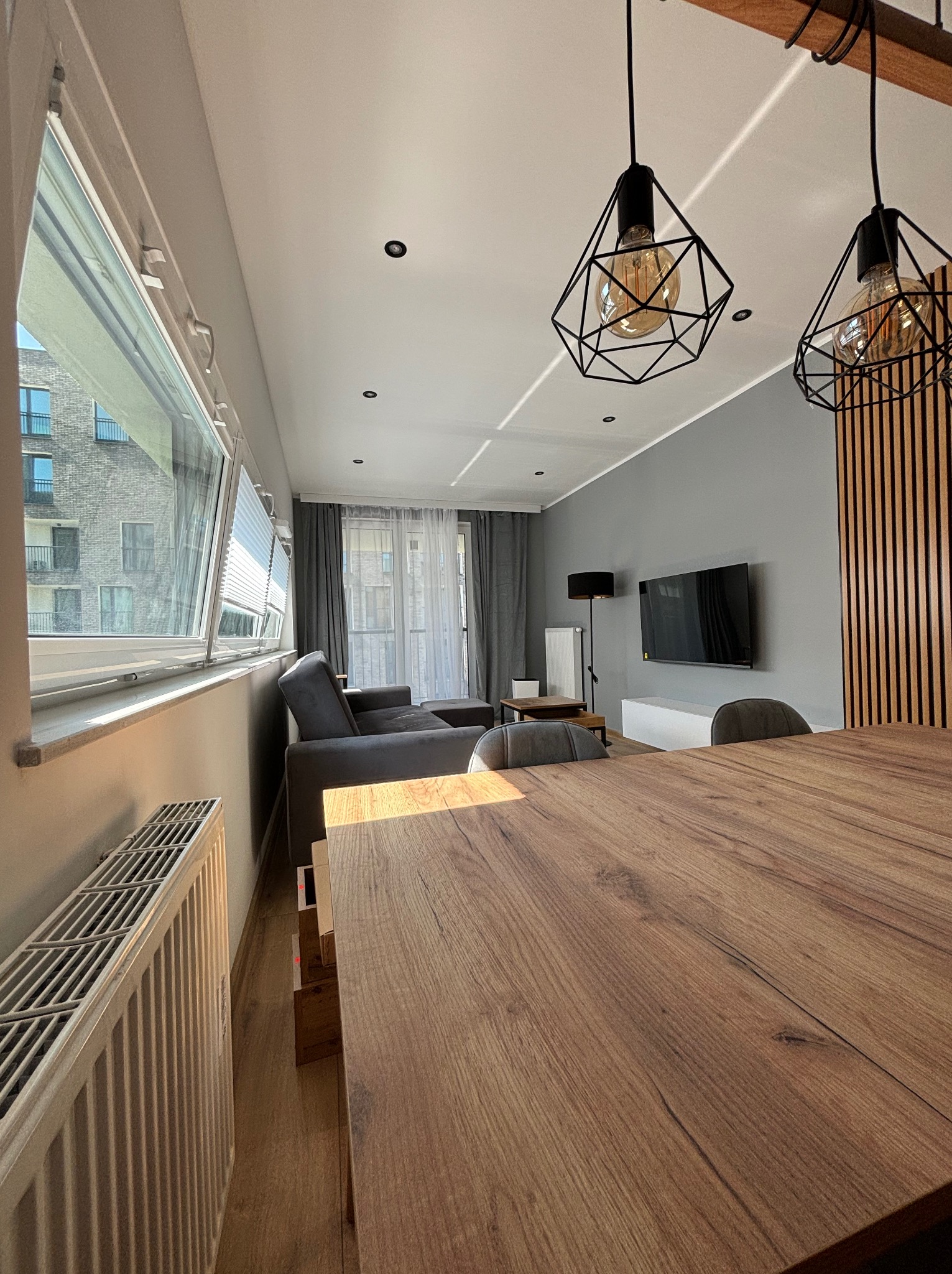Nowoczesny salon z drewnianym stołem na pierwszym planie, szara ściana z telewizorem, geometryczne lampy wiszące i widok na budynek mieszkalny za oknem.