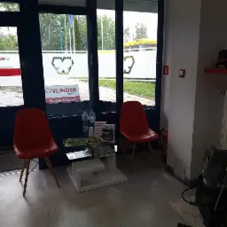 Wnętrze poczekalni firmy z dwoma czerwonymi krzesłami, stolikiem z wodą i przekąskami, widoczne narzędzia na półce i myjka ciśnieniowa.