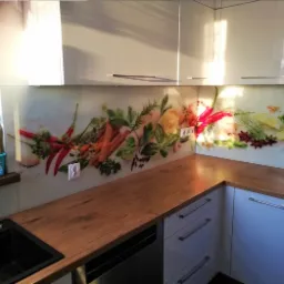 Szklany panel kuchenny z motywem warzyw i przypraw, zamontowany między blatem a szafkami wiszącymi, oświetlony padającym światłem słonecznym.