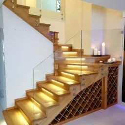 Drewniane schody z podświetleniem LED stopni i szklaną balustradą mocowaną na metalowe uchwyty, zintegrowane z regałem na wino, oświetlone świecami.