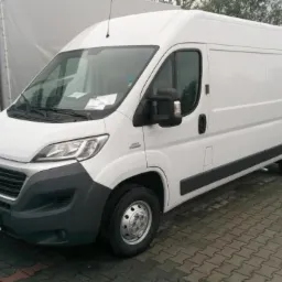 Biały furgon dostawczy Fiat Ducato na kostce brukowej, z widoczną kartką za szybą, zaparkowany obok innego furgonu.