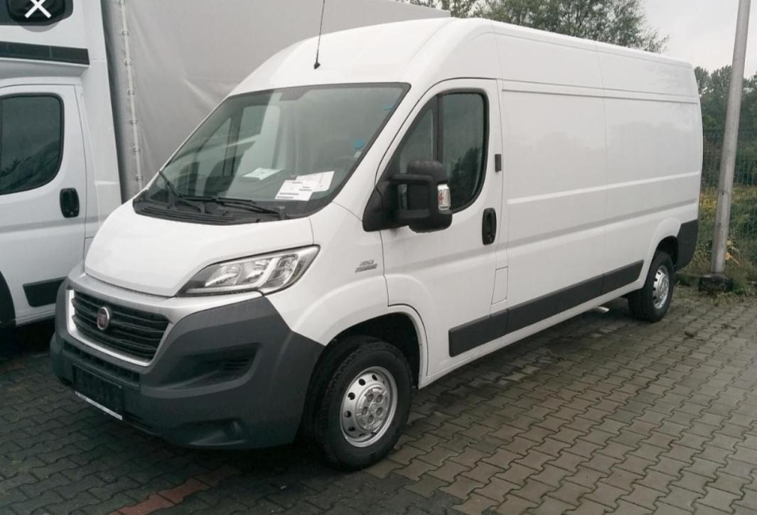 Biały furgon dostawczy Fiat Ducato na kostce brukowej, z widoczną kartką za szybą, zaparkowany obok innego furgonu.