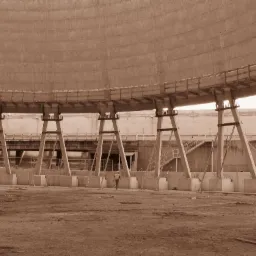 Wnętrze wznoszonej konstrukcji chłodni kominowej, widoczne betonowe filary, stalowe rusztowania i pracownik w oddali, całość utrzymana w sepii.