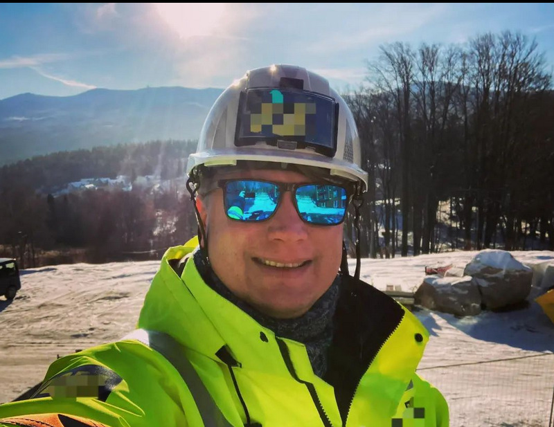 Uśmiechnięty inżynier w kasku i odblaskowej kurtce na tle zimowego krajobrazu z górami. W okularach odbija się budowa. Na kasku zamontowany ekran.