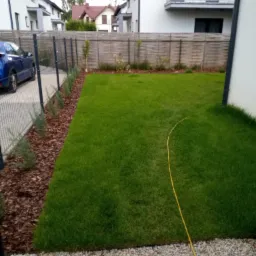 Zadbany trawnik przed nowoczesnym domem z szarą metalową siatką ogrodzeniową, obok której posadzone są małe krzewy wysypane korą. W tle widoczny drewniany płot i zaparkowany niebieski samochód.