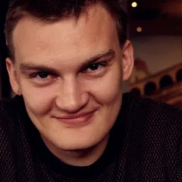 Portret uśmiechniętego mężczyzny w swetrze o drobnej fakturze, z widoczną częścią dekoracji ściennej w tle.