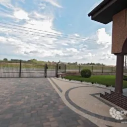 Ułożona z kostki brukowej podjazd i ścieżka prowadzą do bramy wjazdowej i wejścia do domu. Dwukolorowa kostka tworzy wzór łuku. Widoczne jest ogrodzenie posesji i fragment budynku z kolumną.