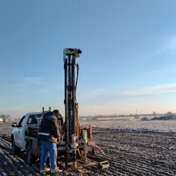 Pracownik obsługuje wiertnicę zamontowaną na samochodzie typu pick-up na zaśnieżonym polu, widoczny numer rejestracyjny PZ 87, w tle zabudowania miejskie.