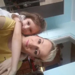 Portret matki z córką, ujęcie z bliska, delikatny makijaż matki, naturalny wyraz twarzy dziecka, jasne tło ściany.