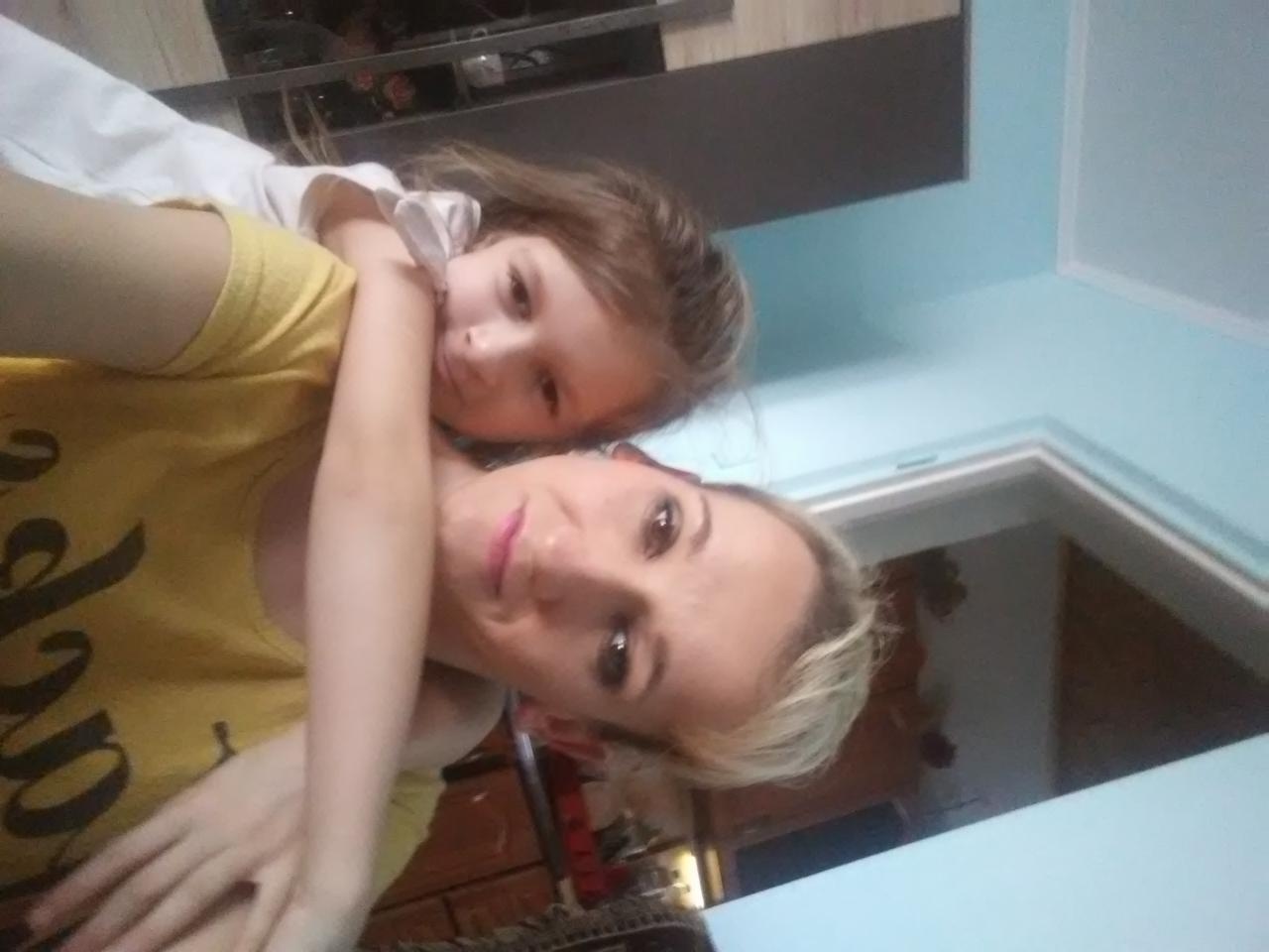 Portret matki z córką, ujęcie z bliska, delikatny makijaż matki, naturalny wyraz twarzy dziecka, jasne tło ściany.