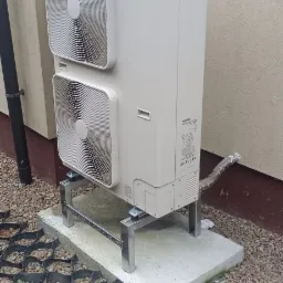 Zewnętrzna jednostka pompy ciepła Toshiba Estia, zamontowana na podwyższeniu z metalowych profili, na betonowej podstawie, obok ściany budynku.