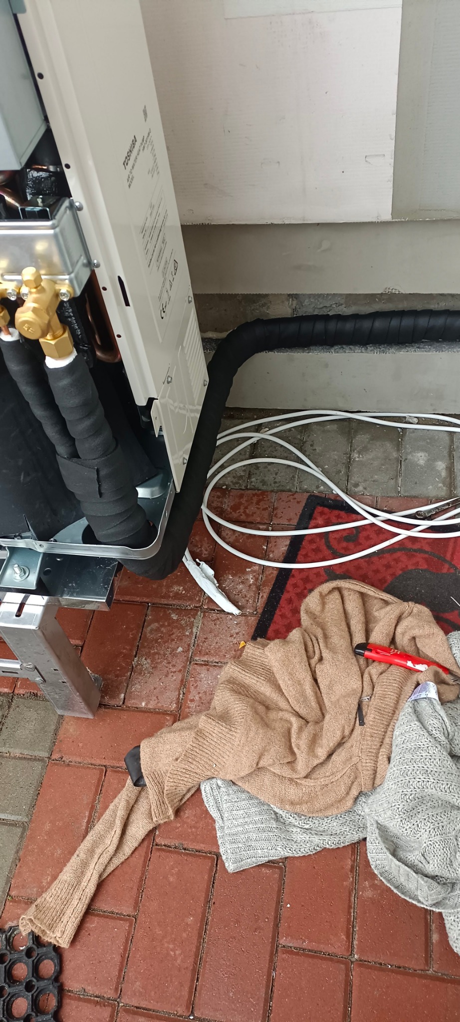 Zewnętrzna jednostka klimatyzacji Toshiba na aluminiowym stelażu, z podłączonymi izolowanymi rurami miedzianymi i przewodami, leżąca na czerwonej ceglanej posadzce, obok której leżą ubrania i nóż...