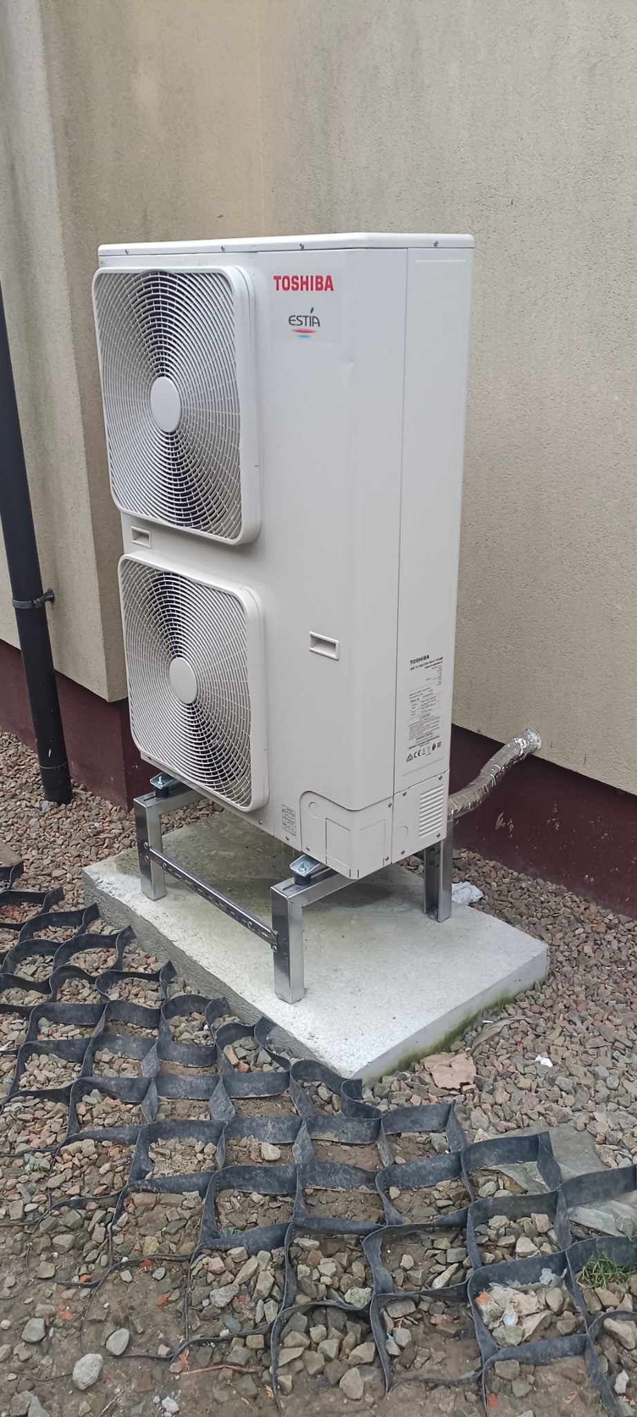 Biała jednostka zewnętrzna pompy ciepła Toshiba Estia, zamontowana na metalowej konstrukcji na betonowej podstawie, widoczna rura w izolacji, tło stanowi elewacja budynku.