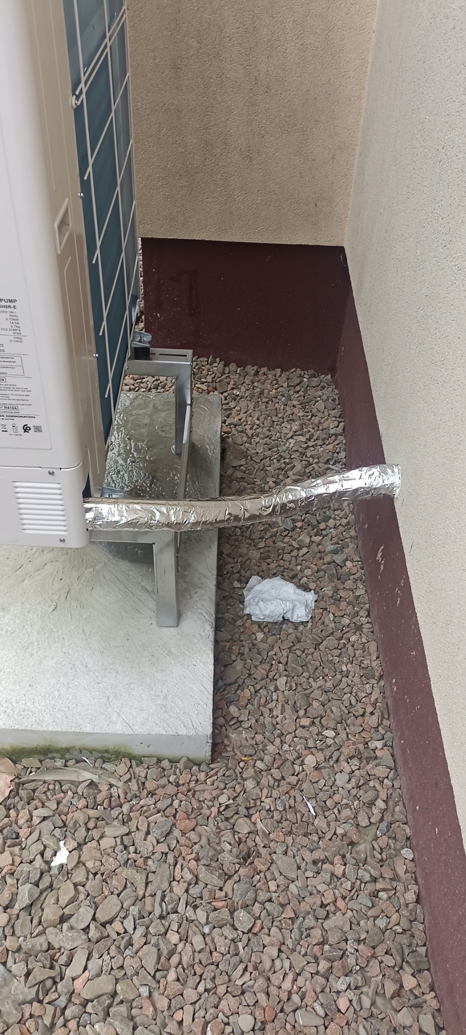 Zewnętrzna jednostka pompy ciepła na betonowej podstawie z izolowanym przewodem odprowadzającym skropliny, otoczona kamieniami i fragmentem ściany.