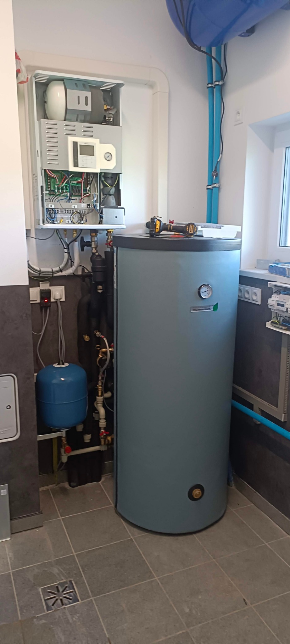 Wnętrze kotłowni z zamontowanym zasobnikiem ciepłej wody użytkowej w kolorze szarym, obok widoczny otwarty panel sterowania kotłem z widocznymi przewodami i elementami elektronicznymi, na zasobniku...