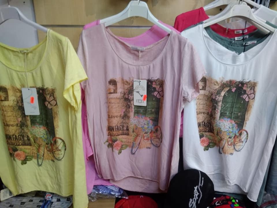 Trzy damskie koszulki z krótkim rękawem na wieszakach: żółta, różowa i biała, każda z nadrukiem przedstawiającym rower z kwiatami i motywem vintage.