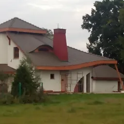 Niedokończony dom jednorodzinny z nietypowym dachem i czerwonym kominem, widoczne rusztowania przy elewacji.