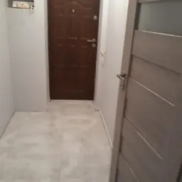 Wnętrze przedpokoju z ułożoną jasną, matową terakotą, widoczne drzwi wejściowe z ciemnego drewna oraz drzwi wewnętrzne w kolorze szarym.