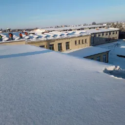 Ekipa w pomarańczowych kombinezonach odśnieża dach budynku szkolnego, widok z góry na zasypany śniegiem dach, błękitne niebo w tle.