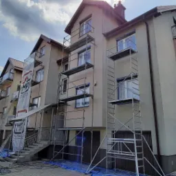 Budynek wielorodzinny w trakcie prac elewacyjnych, widoczne rusztowania, okna zabezpieczone folią, baner reklamowy firmy na rusztowaniu, błękitne płachty zabezpieczające na ziemi.