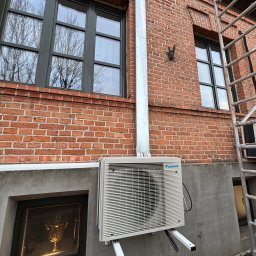 Błysk Paweł Szczepaniak - Zamontowany klimatyzator Daikin na ceglanej ścianie budynku z widoczną rurą odprowadzającą i fragmentem rusztowania. W tle okno z odbiciem drzew.