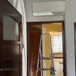 Błysk Paweł Szczepaniak - Wnętrze w trakcie malowania: drabina oparta o ścianę z żółtymi pasami, widoczny klimatyzator i drewniane drzwi. Prace malarskie w toku, widok na okno z firankami.