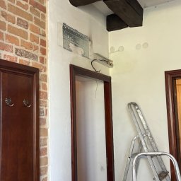 Błysk Paweł Szczepaniak - Wnętrze w trakcie remontu: widoczna ściana z cegły, przygotowane miejsce na klimatyzator, otwory na oświetlenie i drabina oparta o ścianę. Drewniane belki stropowe.