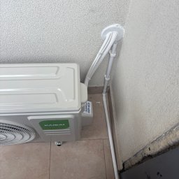 Błysk Paweł Szczepaniak - Klimatyzator Kaisai zamontowany na zewnątrz budynku, z widocznymi przewodami i rurami odprowadzającymi, przymocowanymi do ściany. Widoczny fragment buta w dolnym rogu.