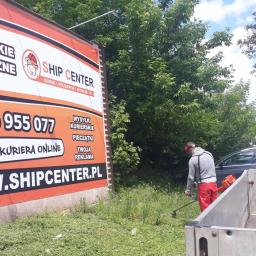 Błysk Paweł Szczepaniak - Pracownik w czerwonych spodniach i szarej bluzie kosi trawę przy użyciu podkaszarki spalinowej przed budynkiem z billboardem reklamowym Ship Center.