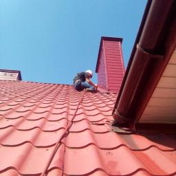 Błysk Paweł Szczepaniak - Pracownik w kasku na czerwonym dachu z blachodachówki, zabezpieczony liną, obok komina z blachy i rynny, na tle błękitnego nieba.